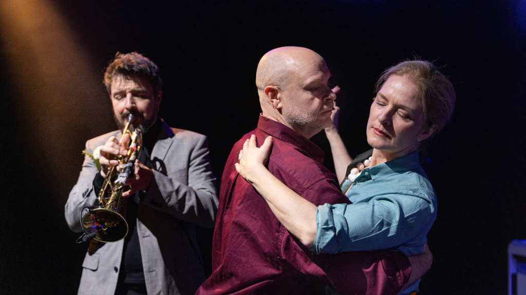 En musiker spelar trumpet i bakgrunden medan en manlig och en kvinnlig skådespelare dansar i förgrunden.