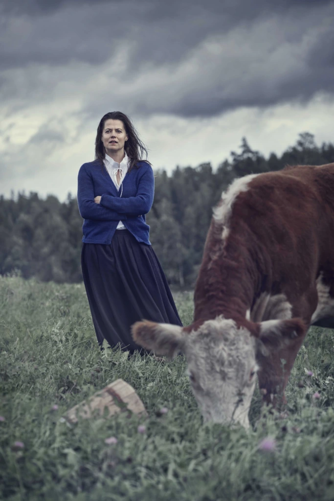 Emma Klingenbergen i rollen som Katrina står med armarna i kors i bakgrunden. I förgrunden betar en ko.