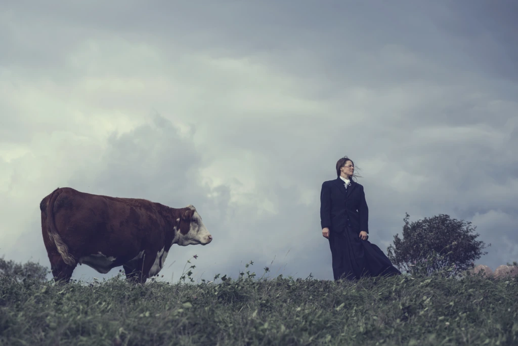 En ko och Emma Klingenberg, som spelar rollen som Katrina står bredvid varandra upp på en grästäckt ås.
