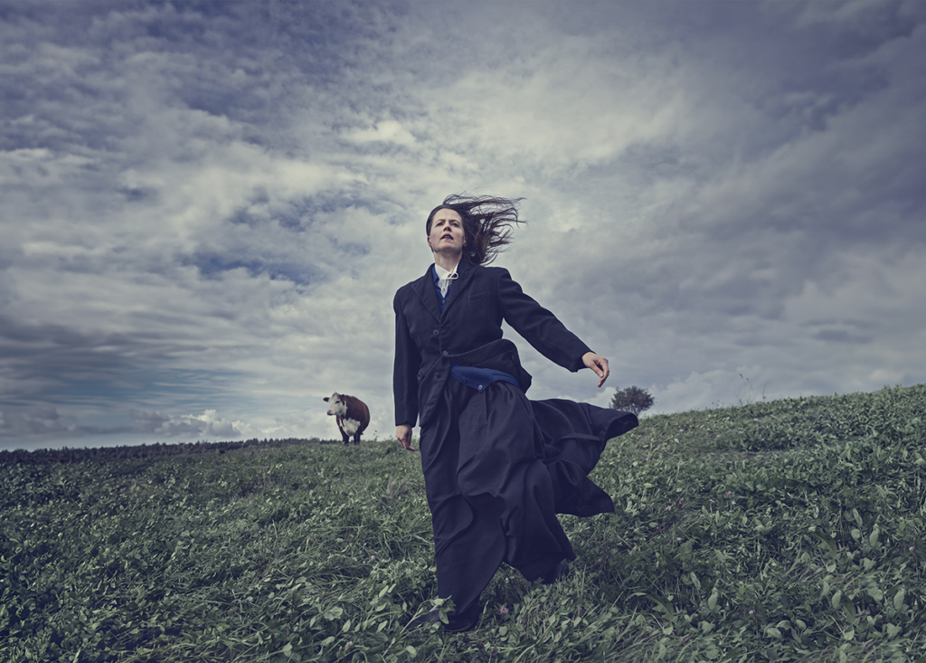 Affischfoto. Katrina (Emma Klingenberg) står på en äng iklädd 1800-talskläder. Vinden blåser i hennes hår och i bakgrunden ser man kor.