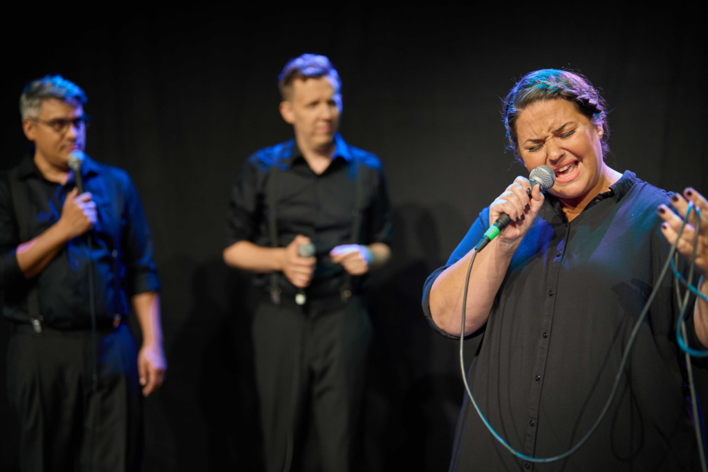 Föreställningsfoto. Daniela Farnzell sjunger i en mikrofon i förgrunden medan Jerry Wahlforss och Samuel Karlsson står i bakgrunden.