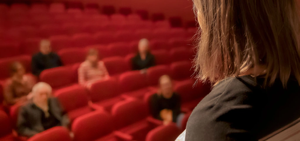 Ett foto av en person som står uppe på scenen och pratar till människor som sitter i Åbo Svenska Teaters salong.