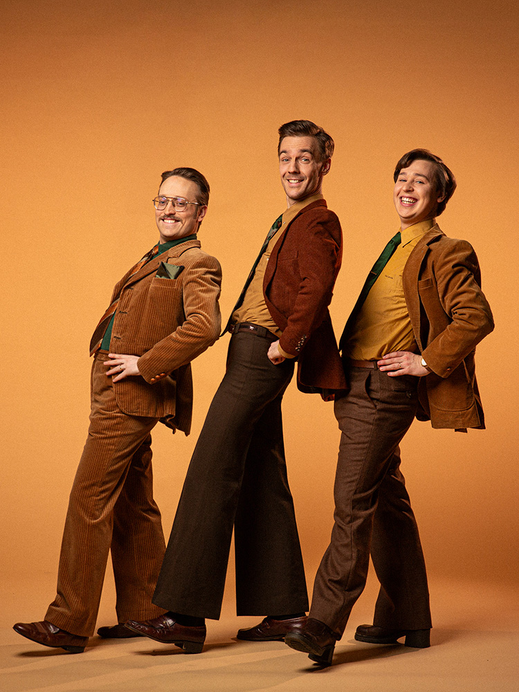 KAJ-trion står uppställda iklädda bruna, 70-talsinspirerade kostymer och slipsar.