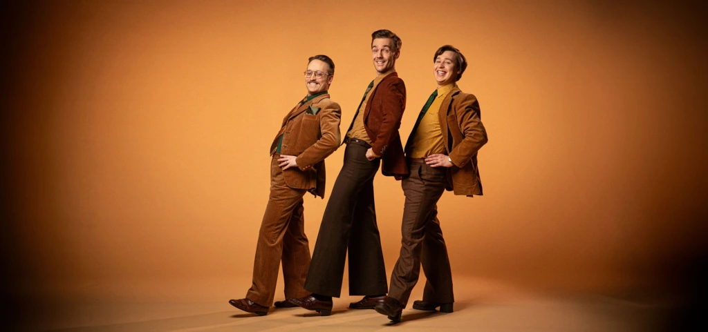KAJ-trion står uppställda iklädda bruna, 70-talsinspirerade kostymer och slipsar.