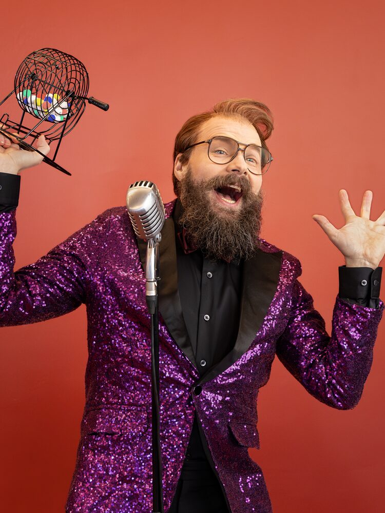 En man i glittrande lila kavaj står vid en mikrofon mot orange bakgrund och håller en bingotrumma med färgglada bollar. Mies kimaltavassa violetissa takissa seisoo mikrofonin äärellä oranssia taustaa vasten ja pitää kädessään bingorumpua. A man in a sparkling purple jacket stands by a microphone against an orange background, holding a bingo cage with colourful balls.