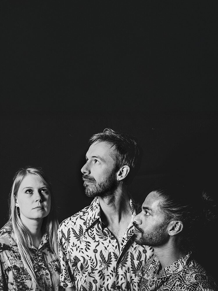 En svartvit studiobild med tre personer som står tätt intill varandra mot en mörk bakgrund. Alla bär mönstrade skjortor och blickar uppåt i olika riktningar med allvarsamma uttryck. Mustavalkoinen studiokuva, jossa kolme henkilöä seisoo lähekkäin tummalla taustalla. Heillä on yllään kuviolliset paidat ja he katsovat eri suuntiin ylöspäin vakavailmeisinä. A black-and-white studio portrait of three people standing close together against a dark background. They are wearing patterned shirts and looking upward in different directions with serious expressions.