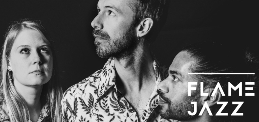 En svartvit närbild av tre personer som ser uppåt åt olika håll, alla klädda i mönstrade skjortor. Bilden har en mörk bakgrund och Flame Jazz-logotypen syns till höger. Mustavalkoinen lähikuva kolmesta henkilöstä, jotka katsovat ylöspäin eri suuntiin kuviollisissa paidoissa. Tausta on tumma ja Flame Jazz -logo näkyy oikealla. A black-and-white close-up of three people looking upward in different directions while wearing patterned shirts. The background is dark and the Flame Jazz logo appears on the right.