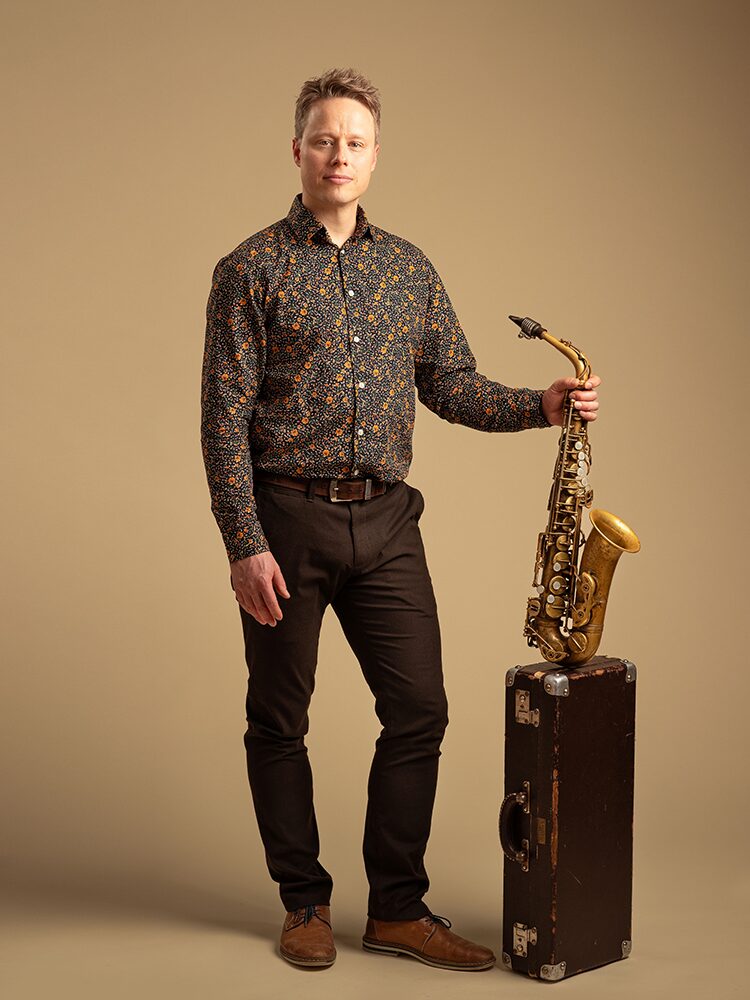 Ett studioporträtt där en person står mot en beige bakgrund och håller en saxofon som vilar på ett mörkt instrumentfodral. Personen bär en mörk, mönstrad skjorta och har ett neutralt uttryck. Studiossa otettu muotokuva, jossa henkilö seisoo beigeä taustaa vasten ja pitää saksofonia, joka lepää tumman instrumenttilaukun päällä. Henkilöllä on tumma, kuviollinen paita ja neutraali ilme. A studio portrait of a person standing against a beige background, holding a saxophone that rests on a dark instrument case. The person wears a dark patterned shirt and has a neutral expression.