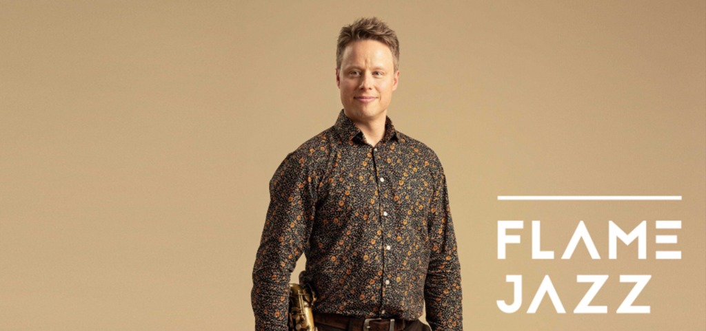 En liggande studiobild av en person mot en beige bakgrund, iklädd en mörk mönstrad skjorta. Personen står bredvid en saxofon och Flame Jazz-logotypen syns till höger. Vaaka-asentoinen studiokuva henkilöstä beigellä taustalla, päällään tumma kuviollinen paita. Henkilön vieressä on saksofoni ja Flame Jazz -logo näkyy oikealla. Vaaka-asentoinen studiokuva henkilöstä beigellä taustalla, päällään tumma kuviollinen paita. Henkilön vieressä on saksofoni ja Flame Jazz -logo näkyy oikealla.