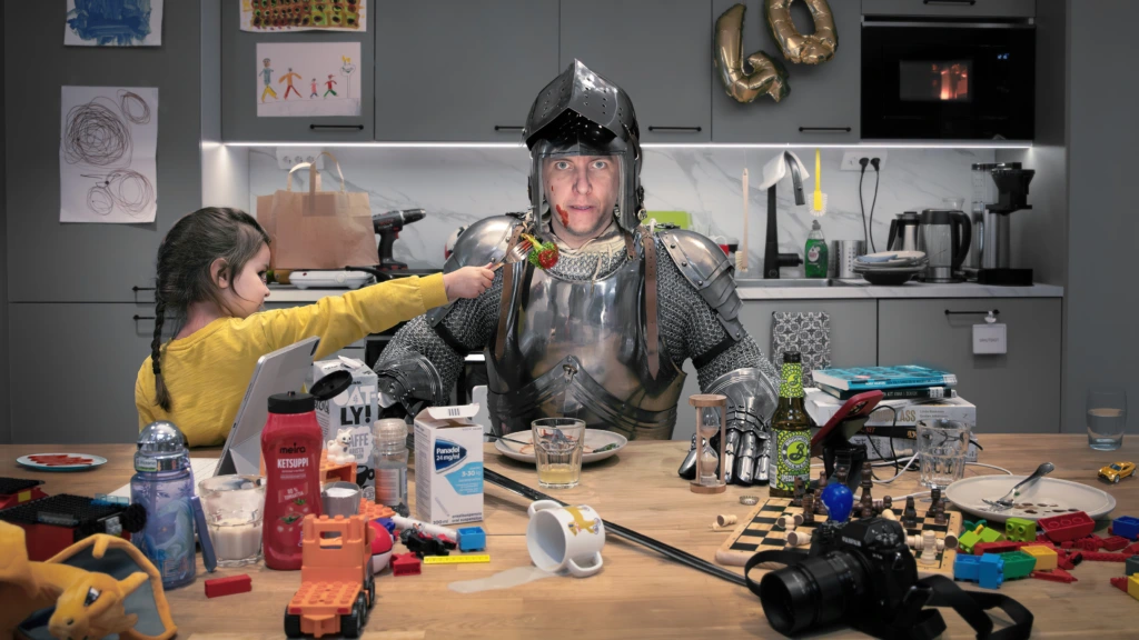 Svenska: En man iklädd en hel rustning sitter vid ett stökigt köksbord fullt av leksaker, muggar, medicinpaket och halvfulla glas. Ett barn håller fram en gaffel med en köttbulle och spaghetti mot honom. På väggen bakom syns ballonger som bildar siffran 40. Reklambild för pjäsen Medeltiden. Finska: Mies, jolla on yllään täysi haarniska, istuu sotkuisen keittiöpöydän ääressä. Pöydällä on leluja, mukeja, lääkepaketteja ja puoliksi täysiä laseja. Lapsi ojentaa hänelle haarukan, jossa on lihapulla ja spagettia. Taustalla näkyy ilmapalloista muodostettu numero 40. Mainoskuva näytelmästä Keskiaika. English: A man wearing a full suit of armor sits at a messy kitchen table filled with toys, mugs, medicine boxes, and half-empty glasses. A child offers him a fork with a meatball and spaghetti. In the background, balloons form the number 40. Promotional image for the play The Middle Ages.