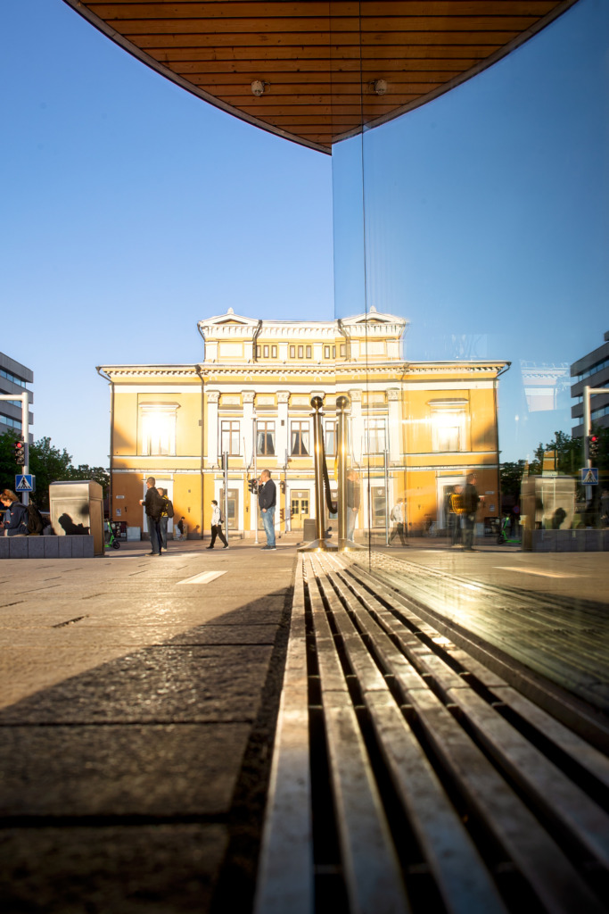 Svenska: Åbo Svenska Teater fotograferad från Salutorget i kvällssol. Flera personer går förbi teaterns gula fasad med vita detaljer, som speglas i glaset från torgets moderna byggnad. Finska: Åbo Svenska Teater kuvattuna Kauppatorilta ilta-auringossa. Useat ihmiset kävelevät ohi keltaisen teatterirakennuksen, jonka valkoiset yksityiskohdat heijastuvat torin modernin rakennuksen lasista. English: Åbo Svenska Teater photographed from the Market Square in the evening sun. Several people walk past the yellow theatre building with white details, reflected in the glass of a modern structure on the square.