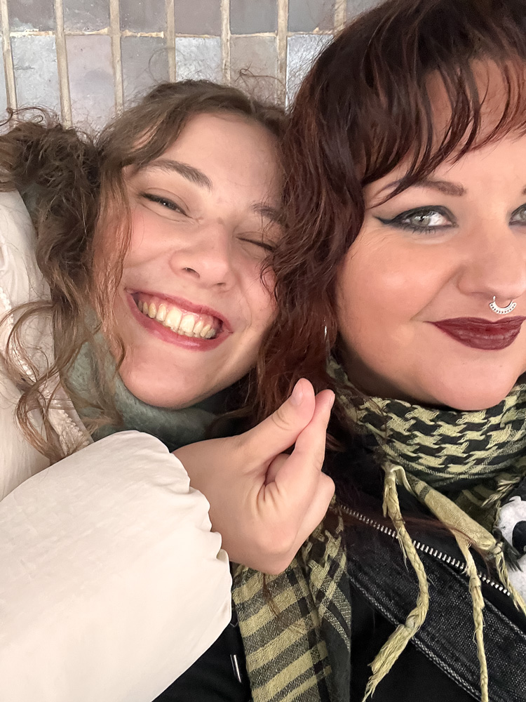 Närbild på två leende personer som lutar kinderna mot varandra. Den ena har lockigt hår och den andra mörkt smink och ett septumpiercing. Lähikuva kahdesta hymyilevästä henkilöstä, jotka nojaavat poskensa toisiaan vasten. Toinen on kiharahiuksinen ja toisella on tummaa meikkiä ja septum-lävistys. Close-up of two smiling people leaning their cheeks together. One has curly hair and the other wears dark makeup and a septum piercing.