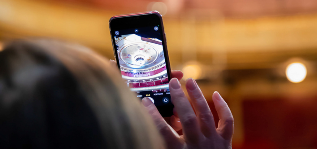En kvinna fotar kristallkronan i salongen. Man ser kronan på telefonskärmen över kvinnan axel.