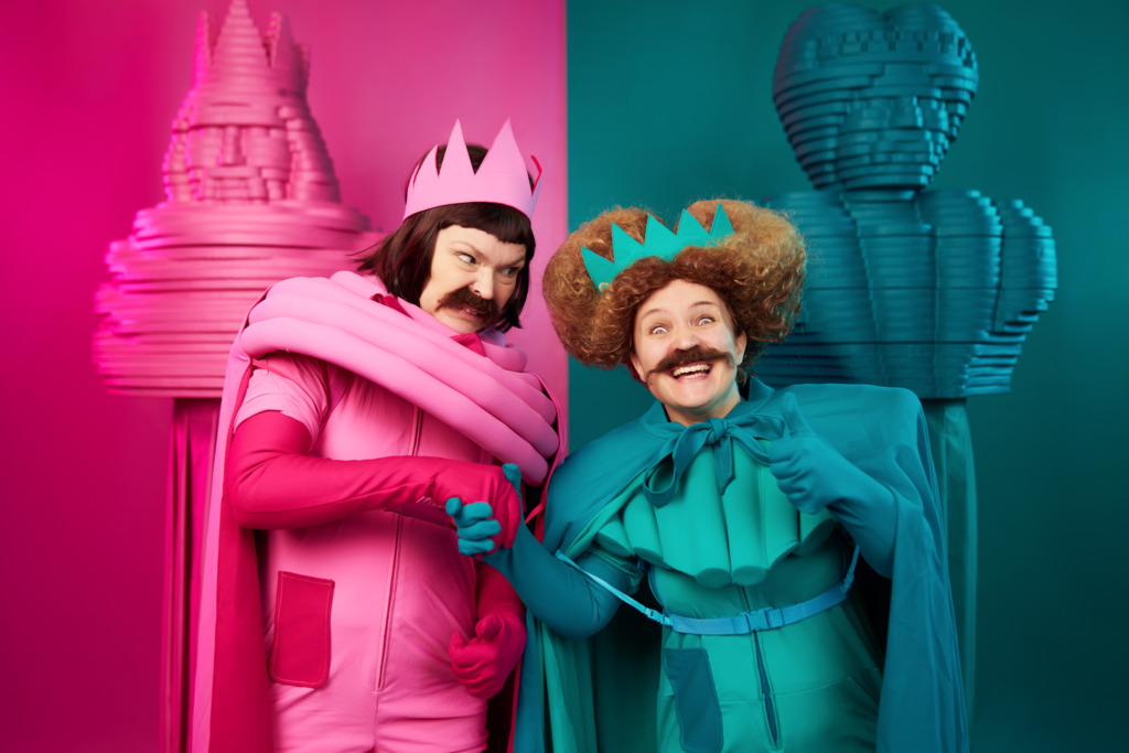SV: Två teaterkaraktärer i starkt rosa och turkos står tätt ihop. De bär kronor, caper och stora mustascher. Den rosa figuren ser misstänksamt på den turkosa, som ler brett. I bakgrunden syns stiliserade skulpturer i samma färgskalor. FI: Kaksi teatterihahmoa kirkkaan pinkissä ja turkoosissa seisoo lähekkäin. Heillä on kruunut, viitat ja suuret viikset. Pinkki hahmo katsoo epäilevästi, turkoosi hymyilee leveästi. Taustalla näkyy samansävyisiä, veistoksellisia muotoja. EN: Two theatrical characters in bright pink and turquoise stand close together. They wear crowns, capes and large moustaches. The pink character looks suspiciously at the turquoise one, who is smiling widely. Stylised sculptures in matching colours appear in the background.