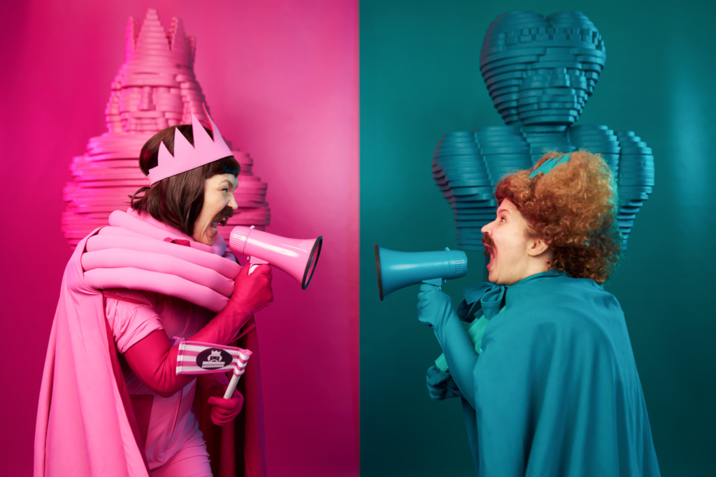 SV: Två färgstarka teaterkaraktärer står mittemot varandra och ropar i varsin megafon. Den rosa figuren till vänster och den turkosa till höger speglar varandras rörelser. Bakgrunden är tydligt uppdelad i rosa och turkos. FI: Kaksi värikästä teatterihahmoa seisoo vastakkain ja huutaa megafoneihin. Vasemmalla pinkki hahmo, oikealla turkoosi, heidän asentonsa peilaavat toisiaan. Tausta jakautuu selkeästi pinkkiin ja turkoosiin. EN: Two colourful theatrical characters face each other, shouting into megaphones. The pink character on the left and the turquoise one on the right mirror each other’s movements. The background is clearly divided into pink and turquoise.