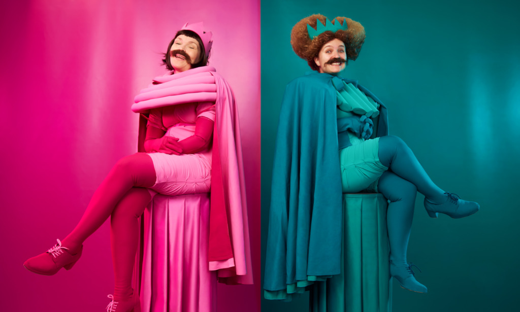 SV: Två teaterkaraktärer sitter självsäkert på varsin piedestal. Den rosa figuren till vänster och den turkosa till höger sitter med korsade ben och ler mot kameran. De bär kronor, caper och mustascher mot en enfärgad bakgrund. FI: Kaksi teatterihahmoa istuu itsevarmasti omilla jalustoillaan. Vasemmalla pinkki hahmo, oikealla turkoosi, molemmat istuvat jalat ristissä ja hymyilevät kameraan. Heillä on kruunut, viitat ja viikset yksiväristä taustaa vasten. EN: Two theatrical characters sit confidently on separate pedestals. The pink character on the left and the turquoise one on the right sit with crossed legs, smiling at the camera. They wear crowns, capes and moustaches against a solid-colour background.