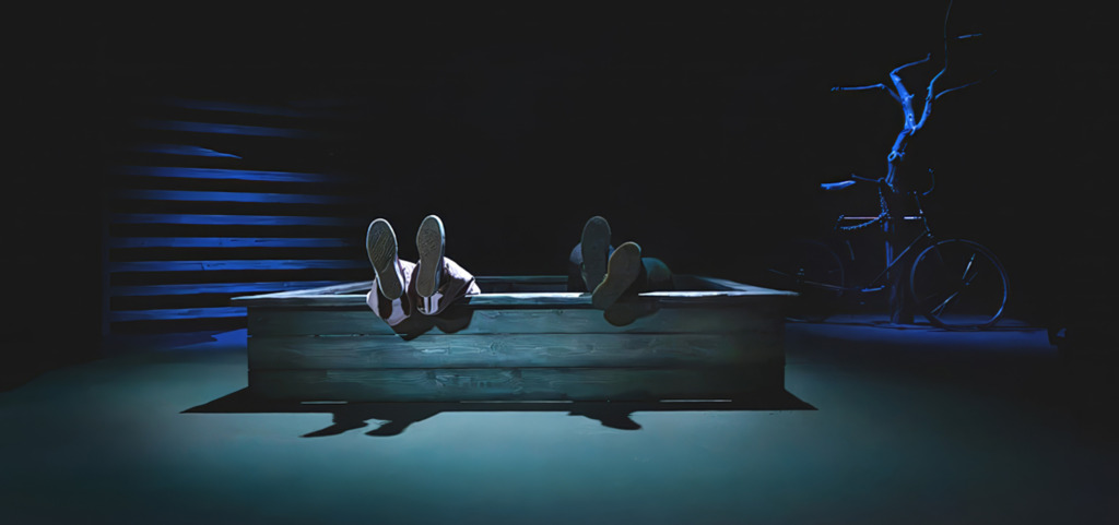 SV: En mörk scenbild där endast två par fötter sticker upp ur en träkonstruktion i mitten av scenen. Blått ljus skapar en drömlik och stillsam stämning. FI: Tumma näyttämökuva, jossa vain kahdet jalat näkyvät puisen rakenteen sisältä keskellä lavaa. Sininen valaistus luo unenomaisen ja hiljaisen tunnelman. EN: A dark stage image where only two pairs of feet are visible sticking out of a wooden structure at centre stage. Blue lighting creates a dreamlike, quiet atmosphere.