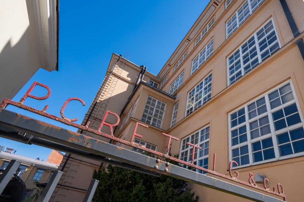 Foto av den ljusgula fasaden till en äldre fabriksbyggnad under klarblå himmel. I förgrunden syns en rostig metallskylt med röda bokstäver som bildar namnet 'P.C. Rettig & Co'. Valokuva vaaleankeltaisesta tehdasrakennuksesta kirkkaan sinisen taivaan alla. Etualalla näkyy ruosteinen metallikyltti, jossa on punaiset kirjaimet 'P.C. Rettig & Co'. Photo of a light yellow industrial building under a clear blue sky. In the foreground, a rusty metal sign with red letters reads 'P.C. Rettig & Co'.