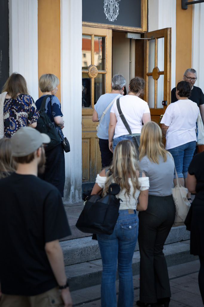 En grupp människor står i kö utanför en byggnad och går in genom en öppen trädörr. Flera personer står på trappsteget, och en person håller upp dörren inifrån. Joukko ihmisiä jonottaa rakennuksen ulkopuolella ja kulkee sisään avoimesta puuovesta. Useita ihmisiä seisoo portailla, ja yksi henkilö pitää ovea auki sisältä käsin. A group of people are standing in a line outside a building, entering through an open wooden door. Several people are on the steps, and one person is holding the door open from inside.