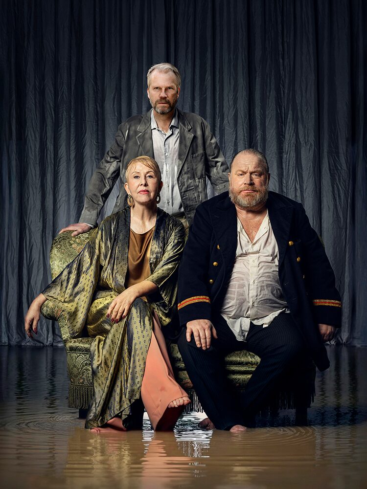 Tre personer poserar i en mörk, dramatisk studio. Två sitter tätt intill varandra i en fåtölj medan den tredje står bakom dem med händerna på ryggstödet. De bär eleganta men slitna scenkläder, och golvet är täckt av grunt vatten som speglar dem. Kolme henkilöä poseeraa tummassa, dramaattisessa studiossa. Kaksi istuu vierekkäin nojatuolissa ja kolmas seisoo heidän takanaan kädet selkänojalla. Heillä on yllään elegantit mutta kulutetut esiintymisvaatteet, ja lattialla oleva matala vesi heijastaa heidän kuvansa. Three people pose in a dark, dramatic studio. Two sit closely together in an armchair while the third stands behind them with hands on the backrest. They wear elegant but worn stage costumes, and the shallow water on the floor reflects their figures.