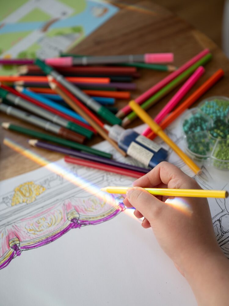 Hand färglägger en teckning med gul penna; färgpennor och pysselmaterial ligger utspridda på bordet. Käsi värittää piirrosta keltaisella kynällä; värikyniä ja askartelutarvikkeita on levällään pöydällä. A hand colors a drawing with a yellow pencil; colored pencils and craft supplies are scattered on the table.