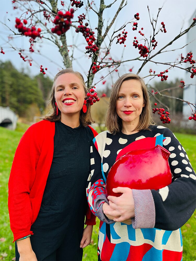 Två personer står bredvid varandra framför ett träd fyllt av röda bär. Den ena bär en röd kofta och svarta kläder, den andra en färgstark tröja i rött, blått, svart och beige och håller en blank röd vas. Bakgrunden består av gräs och träd. Kaksi henkilöä seisoo vierekkäin puun edessä, jossa on punaisia marjoja. Toinen käyttää punaista neuletakkia ja mustia vaatteita, toinen värikästä puna-sinimusta-beigeä neuletta ja pitää käsissään kiiltävää punaista maljakkoa. Taustalla näkyy ruohoa ja puita. Two people stand side by side in front of a tree filled with red berries. One wears a red cardigan over black clothing, while the other wears a colorful sweater in red, blue, black and beige and is holding a shiny red vase. Grass and trees form the background.