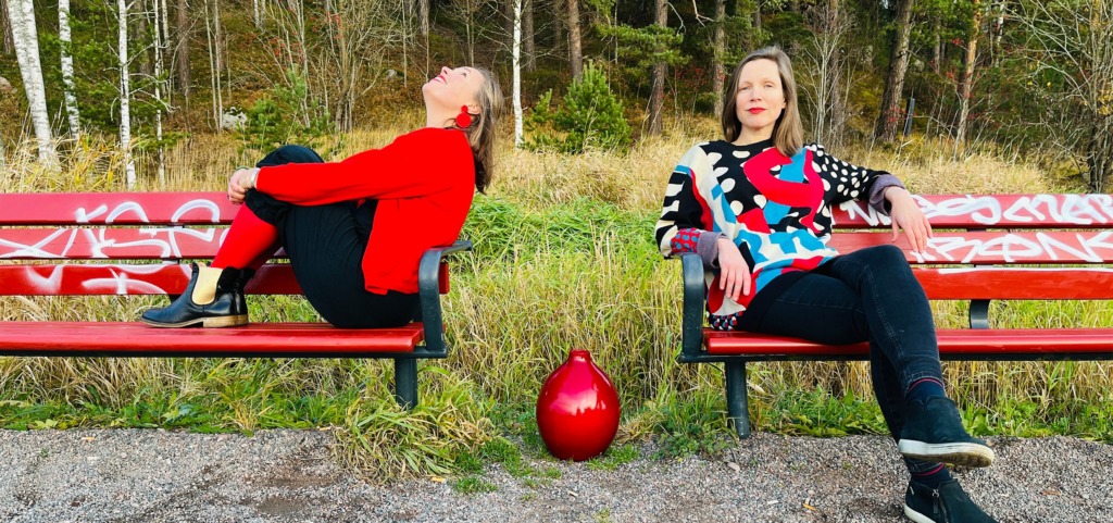 Två personer sitter på varsin röd parkbänk i ett skogsområde. Den ena lutar sig bakåt med benen uppdragna och skrattar, klädd i rött och svart. Den andra sitter avslappnat med benen i kors och bär en färgrik tröja i rött, blått, svart och beige. En blank röd vas står på marken mellan bänkarna. Kaksi henkilöä istuu erillisillä punaisilla puistonpenkeillä metsäisessä ympäristössä. Toinen nojaa taaksepäin jalat koukussa ja nauraa, pukeutuneena punaiseen ja mustaan. Toinen istuu rennosti jalat ristissä ja käyttää värikästä puna-sinimusta-beigeä neuletta. Kiiltävä punainen maljakko on penkkien välissä maassa. Two people sit on separate red park benches in a wooded area. One leans back with legs pulled up and is laughing, dressed in red and black. The other sits relaxed with legs crossed, wearing a colorful sweater in red, blue, black and beige. A shiny red vase is placed on the ground between the benches.