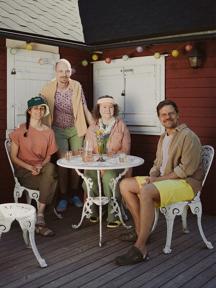 Fyra personer sitter och står kring ett litet vitt trädgårdsbord på en uteveranda framför ett rött hus. De är avslappnat klädda i pastell- och jordnära färger. På bordet står glas och en vas med sommarblommor. Färgglada ljusslingor hänger ovanför dem. Neljän hengen seurue istuu ja seisoo pienen valkoisen puutarhapöydän ympärillä verannalla punaisen rakennuksen edessä. He ovat rennosti pukeutuneita pastelli- ja maanläheisiin sävyihin. Pöydällä on laseja ja maljakko kesäkukilla. Yläpuolella roikkuu värikäs valosarja. Four people sit and stand around a small white garden table on a porch in front of a red house. They are casually dressed in pastel and earthy tones. On the table are glasses and a vase with summer flowers. A string of colorful lights hangs above them.