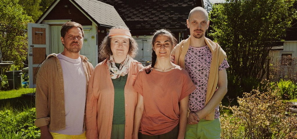 Fyra personer står på rad i en solig trädgård framför ett rött hus med vita detaljer. De bär kläder i mjuka pastellfärger och ser lugna och avslappnade ut. Grönska och buskar omger dem i förgrunden. Neljän hengen ryhmä seisoo riveissä aurinkoisessa puutarhassa punaisen talon edessä. Heillä on yllään pehmeän pastellisia vaatteita ja he näyttävät rauhallisilta ja rennolta. Edustalla on vihreää kasvillisuutta ja pensaita. Four people stand in a row in a sunny garden in front of a red house with white trim. They wear soft pastel-colored clothing and appear calm and relaxed. Greenery and bushes fill the foreground.