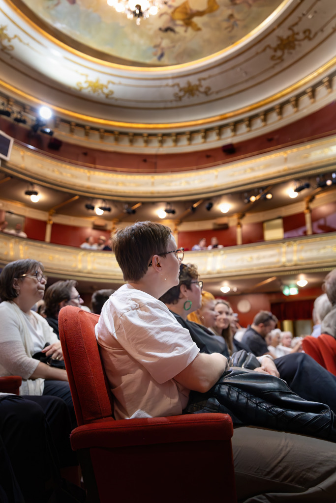 En publik sitter i röda plyschstolar i Åbo Svenska Teaters salong, fotograferad från sidan. Fokus ligger på en person med kort hår och glasögon i förgrunden, medan den rikt utsmyckade takmålningen och de gyllene balkongerna syns ovanför. Stämningen är förväntansfull inför föreställningen. Yleisö istuu punaisilla plyysituoleilla Åbo Svenska Teaterin salissa, kuvattuna sivulta. Etualalla on lyhyttukkainen henkilö, jolla on silmälasit, ja ylhäällä näkyvät teatterin koristeellinen kattomaalaus ja kultaiset parvet. Tunnelma on odottava ennen esitystä. An audience sits in red velvet seats in the auditorium of Åbo Svenska Teater, photographed from the side. The focus is on a short-haired person wearing glasses in the foreground, while the ornate ceiling painting and golden balconies are visible above. The atmosphere is filled with anticipation before the performance.