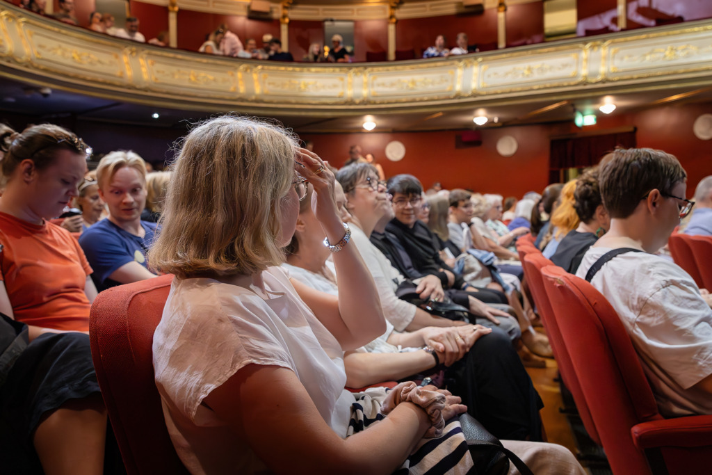 En publik sitter i röda plyschstolar i Åbo Svenska Teaters salong, fotograferad från sidan. Fokus ligger på en person med kort hår och glasögon i förgrunden, medan den rikt utsmyckade takmålningen och de gyllene balkongerna syns ovanför. Stämningen är förväntansfull inför föreställningen. Yleisö istuu punaisilla plyysituoleilla Åbo Svenska Teaterin salissa, kuvattuna sivulta. Etualalla on lyhyttukkainen henkilö, jolla on silmälasit, ja ylhäällä näkyvät teatterin koristeellinen kattomaalaus ja kultaiset parvet. Tunnelma on odottava ennen esitystä. An audience sits in red velvet seats in the auditorium of Åbo Svenska Teater, photographed from the side. The focus is on a short-haired person wearing glasses in the foreground, while the ornate ceiling painting and golden balconies are visible above. The atmosphere is filled with anticipation before the performance.
