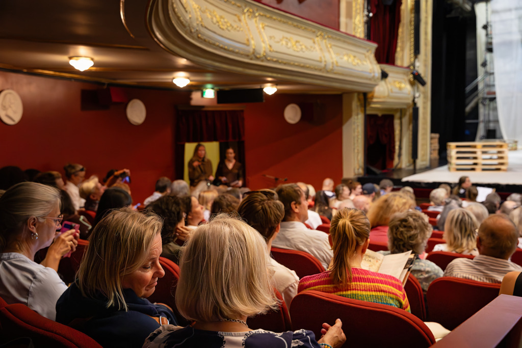 En publik sitter tätt i de röda salongsstolarna i Åbo Svenska Teater. Den rikt dekorerade balkongen med gulddetaljer syns ovanför, och människor i olika åldrar samtalar och bläddrar i programblad inför en föreställning. Yleisö istuu tiiviisti Åbo Svenska Teatterin punaisilla katsomopenkeillä. Yläpuolella näkyy runsaasti koristeltu, kultakoristeinen parveke, ja eri-ikäiset katsojat keskustelevat ja selaavat käsiohjelmia ennen esityksen alkua. An audience is seated closely together in the red auditorium seats at Åbo Svenska Teater. Above them, a richly decorated balcony with gold details is visible, while people of different ages chat and read programmes before the performance begins.