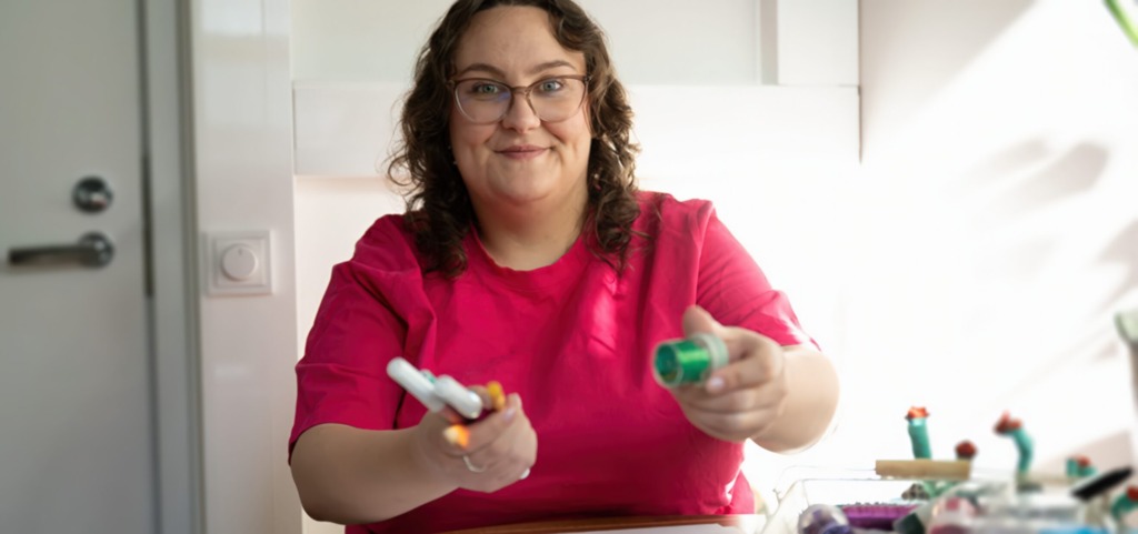 Person i rosa tröja sitter vid pysselbord och räcker fram pysselmaterial i en ljus inomhusmiljö. Pinkkipaitainen henkilö istuu askartelupöydän ääressä ja ojentaa askartelutarvikkeita valoisassa sisätilassa. A person in a pink top sits at a craft table and offers craft supplies in a bright indoor setting.