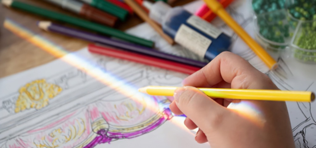 Närbild av hand som färglägger en teckning; pennor och material ligger i bakgrunden. Lähikuva kädestä värittämässä piirrosta; kyniä ja tarvikkeita näkyy taustalla. Close-up of a hand coloring a drawing; pens and materials are visible in the background.