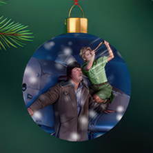 Ett runt julgransornament i glas som visar en dramatisk scen där en pojke svävar i luften medan en man sträcker sig upp mot honom, omgivna av ett blåtonat sken och snö.
Pyöreä lasinen joulukuusikoriste, jossa on dramaattinen kohtaus: poika leijuu ilmassa samalla kun mies kurottaa häntä kohti, ympärillään sinertävä valo ja lumisade.
A round glass Christmas ornament depicting a dramatic moment where a boy floats in the air as a man reaches up toward him, surrounded by a blue glow and falling snow.