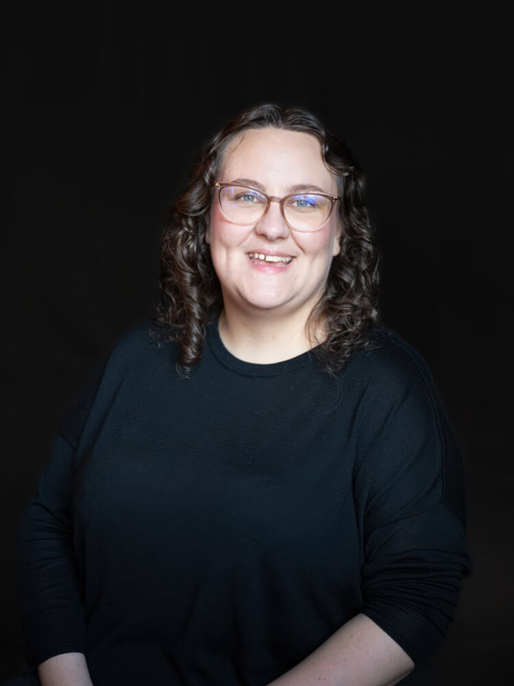 Porträtt av leende person med glasögon i svart tröja mot mörk bakgrund. Muotokuva hymyilevästä silmälasipäisestä henkilöstä mustassa paidassa tummaa taustaa vasten. Portrait of a smiling person with glasses in a black top against a dark background.