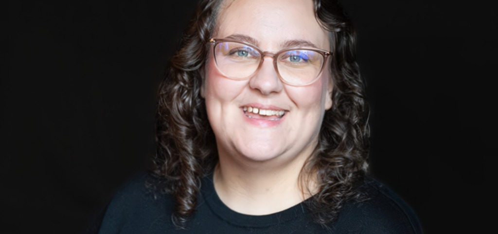 Närbild av leende person med glasögon mot mörk bakgrund. Lähikuva hymyilevästä silmälasipäisestä henkilöstä tummaa taustaa vasten. Close-up of a smiling person with glasses against a dark background.