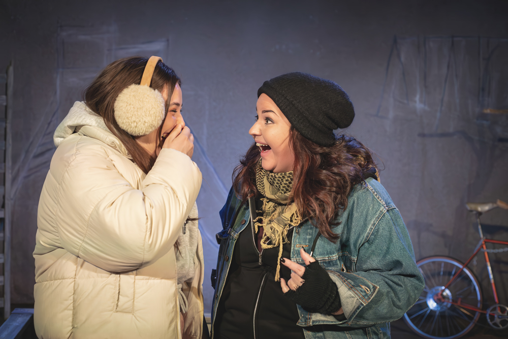 SV: Två unga kvinnor står tätt intill varandra. Den ena, klädd i vit vinterjacka och öronmuffar, skrattar och håller handen för munnen. Den andra, i mössa, jeansjacka och halsduk, ser glatt och överraskat på henne. FI: Kaksi nuorta naista seisoo lähekkäin. Toinen, valkoisessa talvitakissa ja korvaläpissä, nauraa käsi suun edessä. Toinen, pipo päässä, farkkutakissa ja huivissa, katsoo häntä iloisesti ja yllättyneenä. EN: Two young women stand close together. One, wearing a white winter jacket and earmuffs, laughs with her hand over her mouth. The other, in a beanie, denim jacket and scarf, looks at her with a joyful, surprised expression.