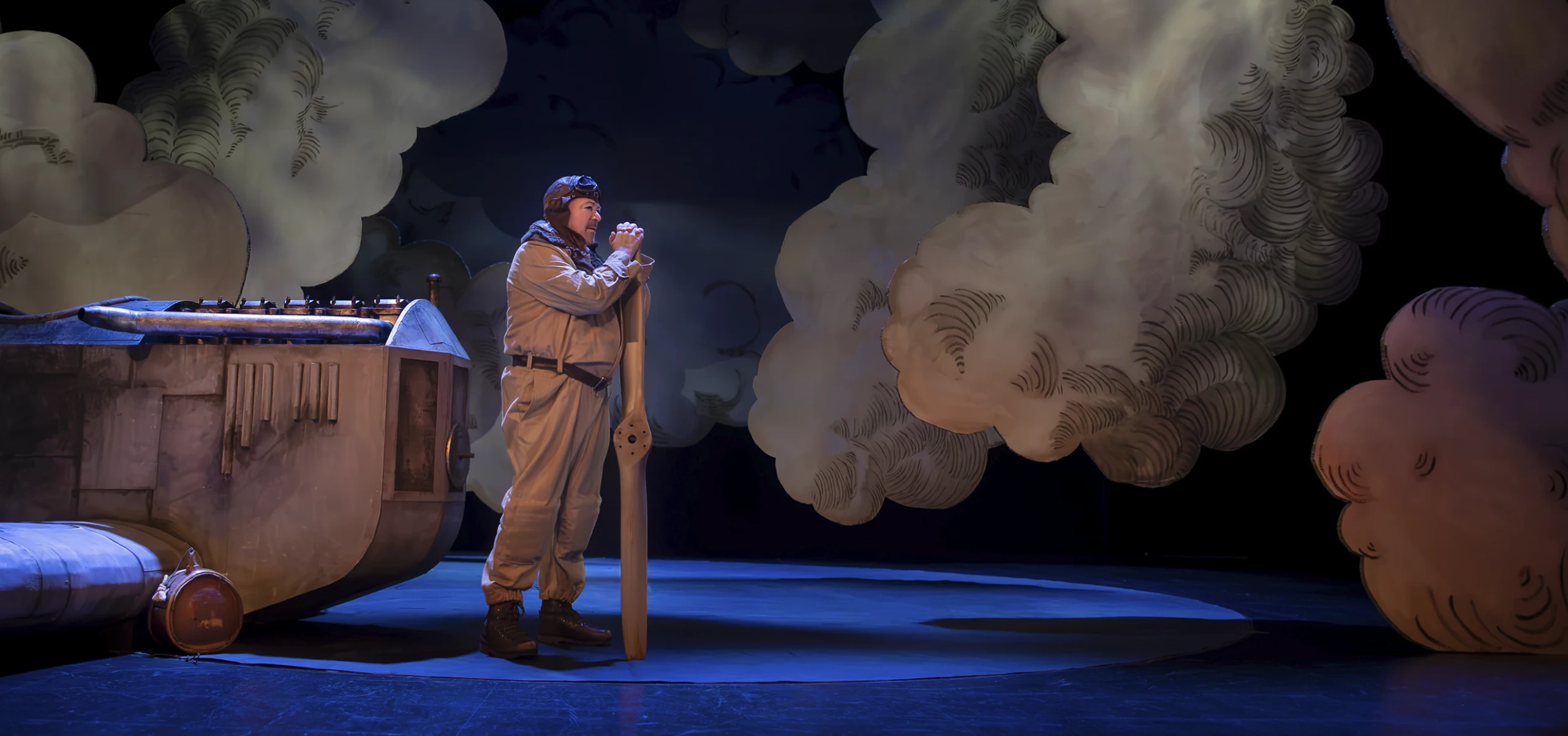 En pilot står på scen bredvid ett litet flygplan och håller i en propeller, omgiven av stora stiliserade moln. Lentäjä seisoo näyttämöllä pienen lentokoneen vieressä ja pitää potkurista kiinni suurten tyyliteltyjen pilvien ympäröimänä. A pilot stands on stage beside a small airplane holding a propeller, surrounded by large stylized clouds.