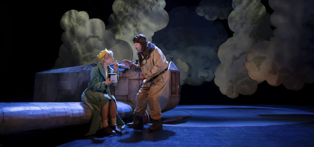 Den lille prinsen sitter på flygplanet med sin krona och en kopp i handen medan piloten talar med honom. Pikku prinssi istuu lentokoneen päällä kruunu päässään ja kuppi kädessään, kun lentäjä puhuu hänelle. The Little Prince sits on the airplane wearing his crown and holding a cup while the pilot speaks to him.