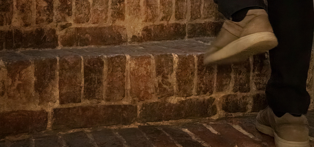 Närbild av fötter i skor som går uppför slitna tegeltrappor inomhus. Lähikuva kengissä olevista jaloista, jotka nousevat kuluneita tiiliportaita sisätiloissa. Close-up of feet wearing shoes walking up worn brick steps indoors.