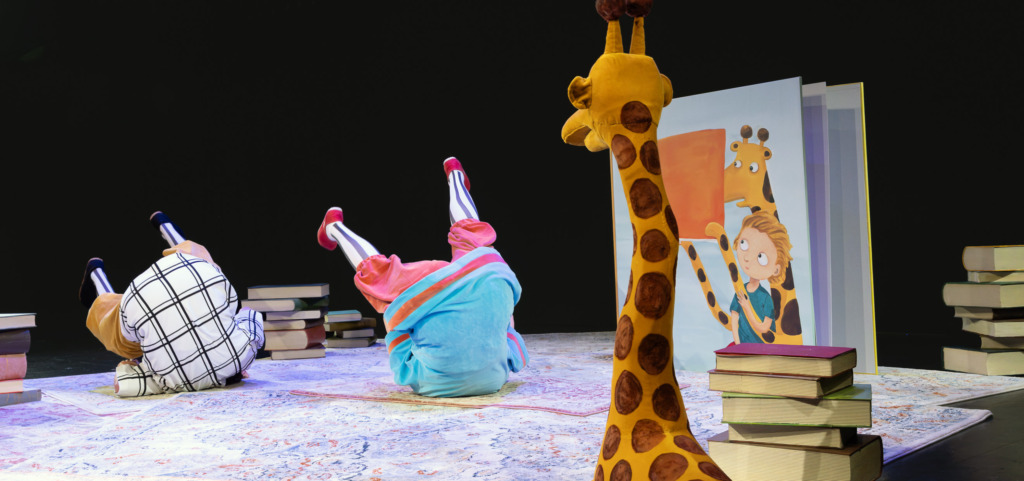 Två skådespelare gör kullerbyttor på scenen bland bokhögar, en stor bilderbok och en giraff. Kaksi näyttelijää tekee kuperkeikkoja näyttämöllä kirjapinojen, suuren kuvakirjan ja kirahvin keskellä. Two actors tumble across the stage among stacks of books, a giant picture book and a giraffe.