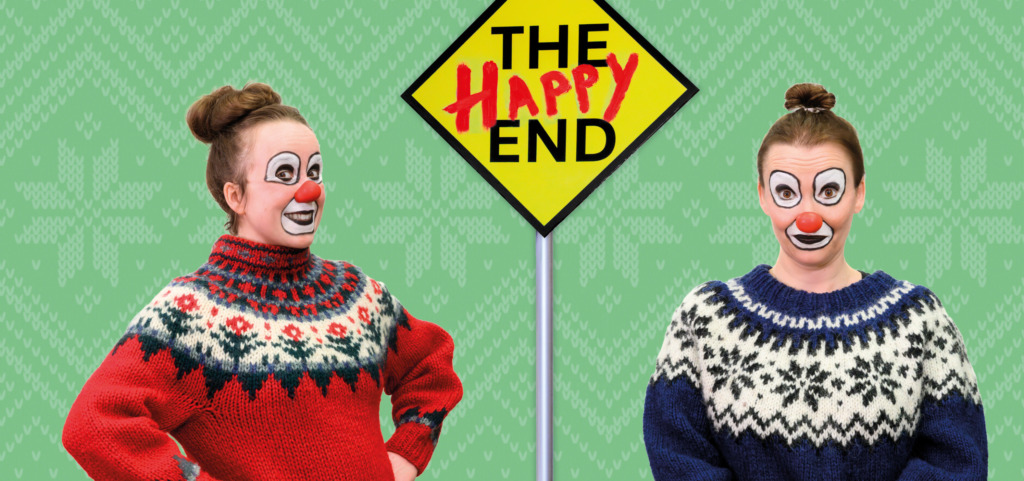 Två clowner i stickade tröjor framför en gul skylt med texten ”The Happy End”. Kaksi klovnia neulepuseroissa keltaisen ”The Happy End” -kyltin edessä. Two clowns in knitted sweaters in front of a yellow sign reading “The Happy End”.
