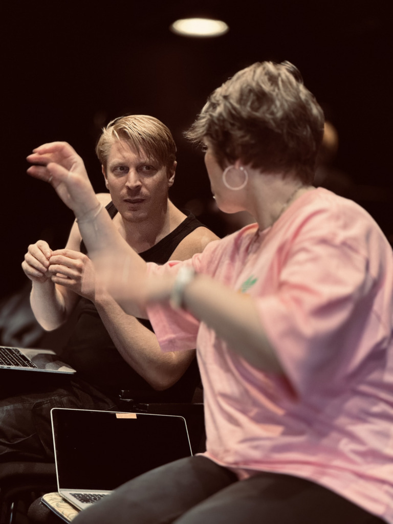 Två personer diskuterar en scen under repetition, med bärbara datorer framför sig.
Kaksi henkilöä keskustelee kohtauksesta harjoituksissa, kannettavat tietokoneet edessään.
Two people discuss a scene during rehearsal, with laptops in front of them.