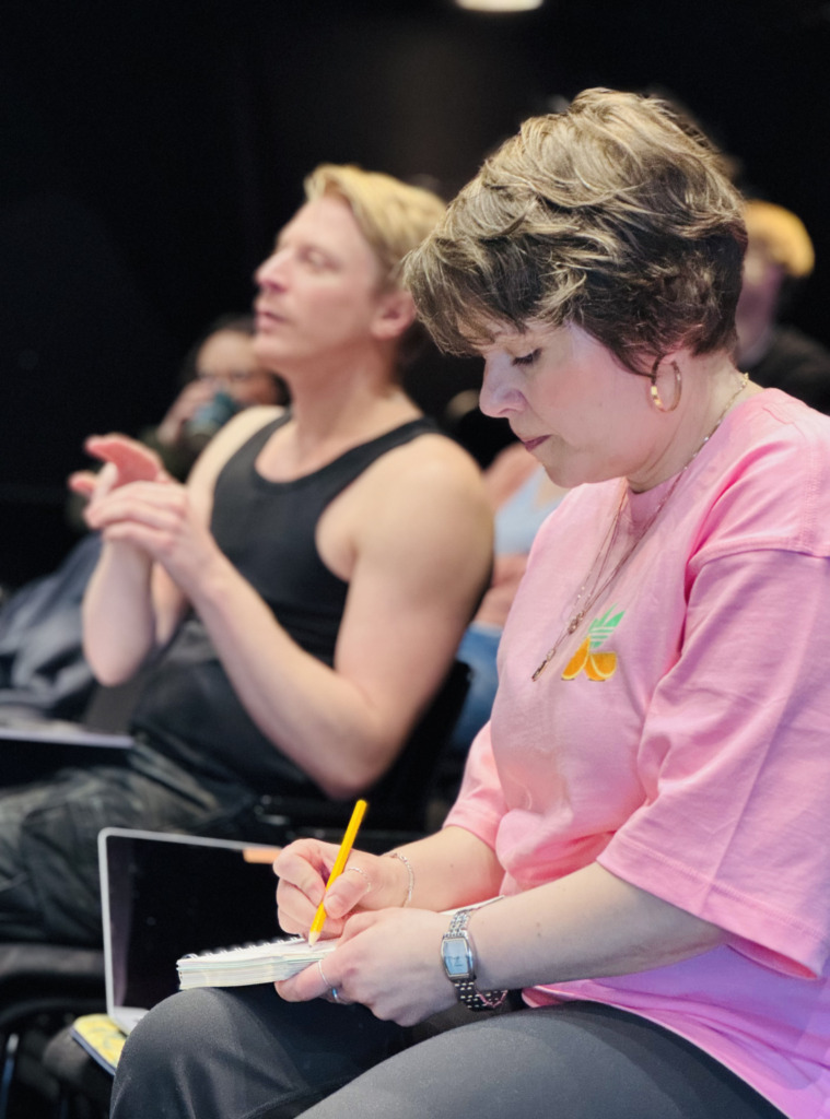 Person i rosa tröja antecknar under teaterrepetition medan andra deltagare sitter i bakgrunden. Vaaleanpunaisessa paidassa oleva henkilö tekee muistiinpanoja teatteriharjoituksissa muiden osallistuessa taustalla. Person in a pink shirt takes notes during a theatre rehearsal while others sit in the background.