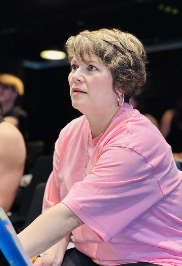 Person i rosa tröja lyssnar uppmärksamt under repetition i teatersalongen. Vaaleanpunaisessa paidassa oleva henkilö kuuntelee tarkkaavaisesti teatteriharjoituksissa. Person in a pink shirt listens attentively during a rehearsal in the theatre auditorium.